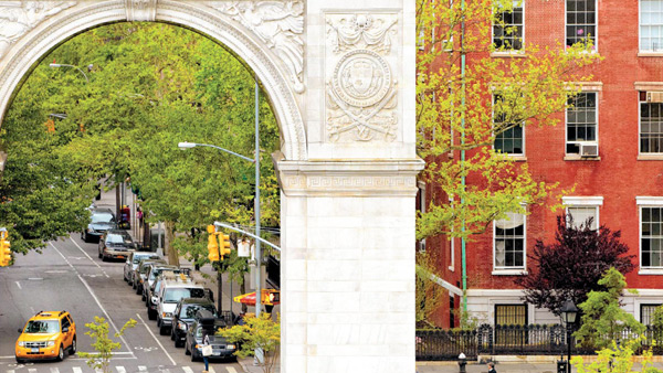 The Washington Square Arch on the left and a red brick building on the right, with trees, parked cars, and a taxi in the background on a street in Greenwich Village, downtown Manhattan, New York City.