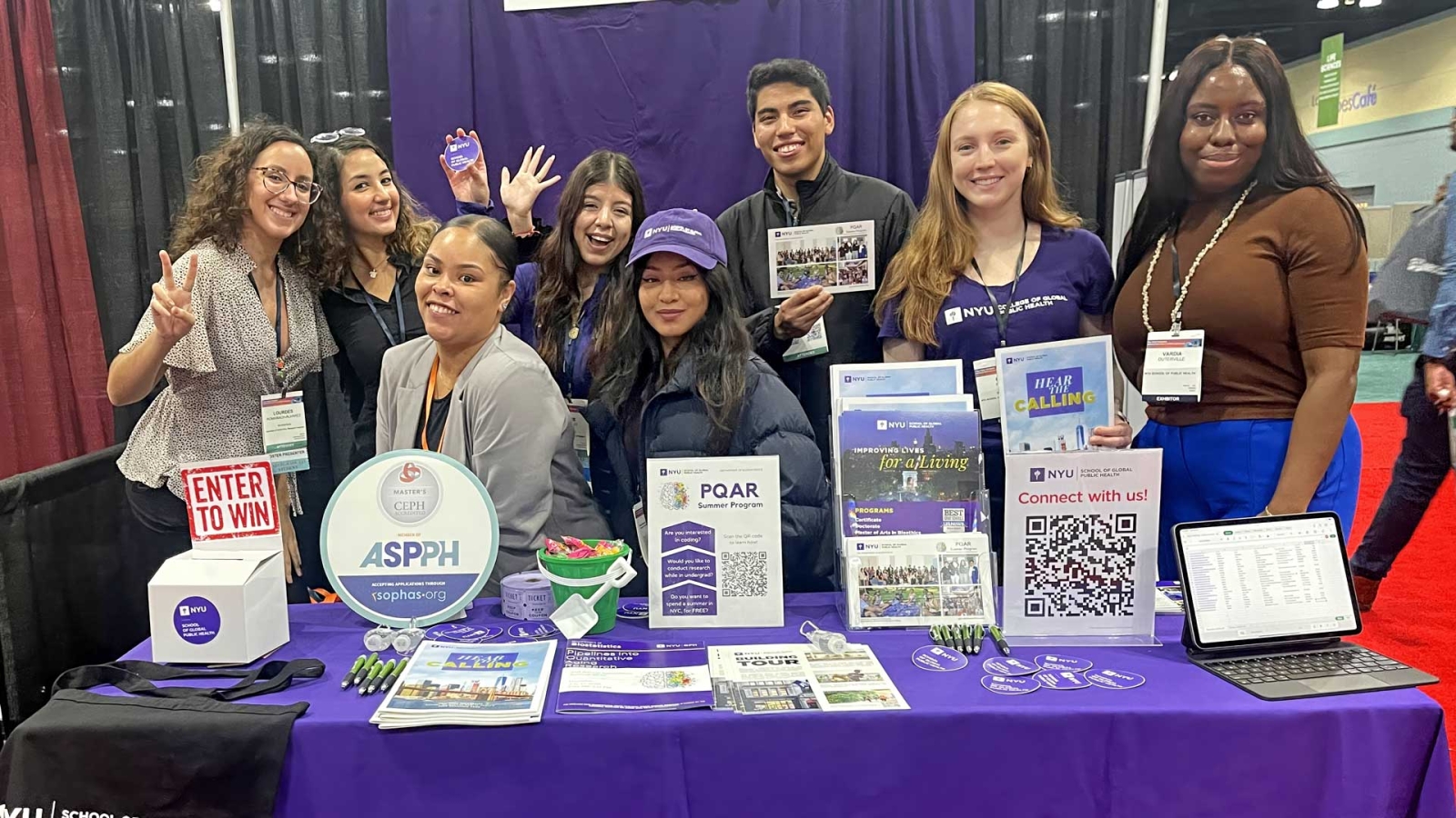 NYU GPH Master of Arts in Bioethics students pose behind an NYU table displaying informational materials and promotional items.